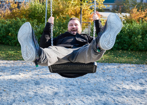 volwassen bebaarde man rijdt een kind swing en lacht - Foto, afbeelding