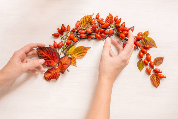 Жіночі руки роблять осінній вінок з рожевих стегон і червоного осіннього листя на тлі дерев'яної текстури. Декор будинку ручної роботи. Майстер-клас з виготовлення декору. Планшет. Вид зверху
 - Фото, зображення