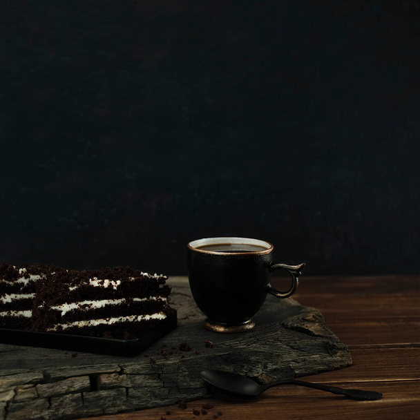 Cup of black coffee with pieces of chocolate cake and spoon on a wooden cracked oak cut. Top view with copy space of dark blue bokeh background - Photo, image