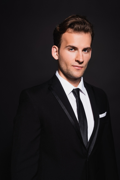 young confident man in elegant suit looking at camera on black - Foto, afbeelding