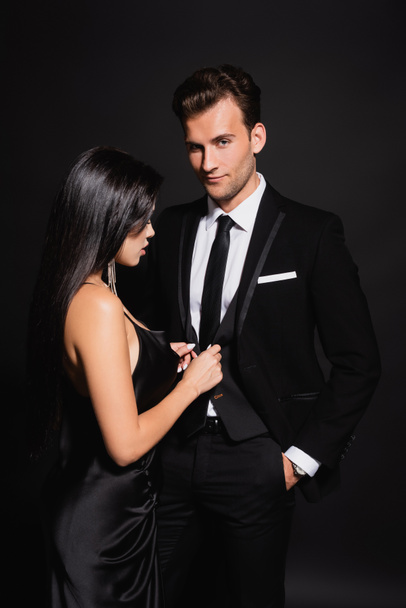 sensual brunette woman unbuttoning vest of elegant man standing with hand in pocket on black - 写真・画像