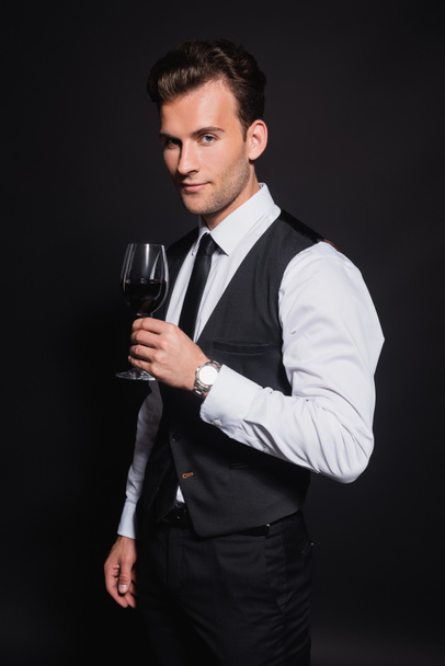 positive and confident man looking at camera while holding glass of red wine isolated on black - Zdjęcie, obraz