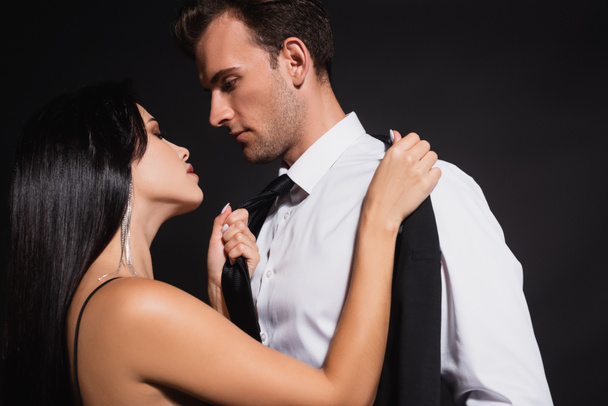 passionate brunette woman pulling tie of man while undressing him isolated on black - Photo, Image