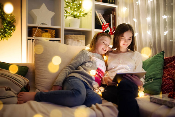 Two cute young sisters using a tablet pc at home in warm and cozy living room at Christmas. Family having online video call on Xmas eve. Winter evening at home with family and kids. - Фото, изображение