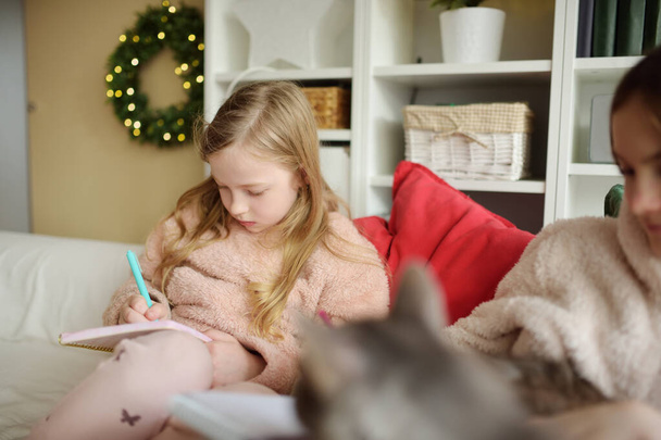 2人のかわいい妹はクリスマスの居心地の良いリビングルームでソファで勉強したりスケッチしたりしました。かわいい子供たちは冬休みに家で時間を過ごす。自宅でクリスマスを祝う. - 写真・画像