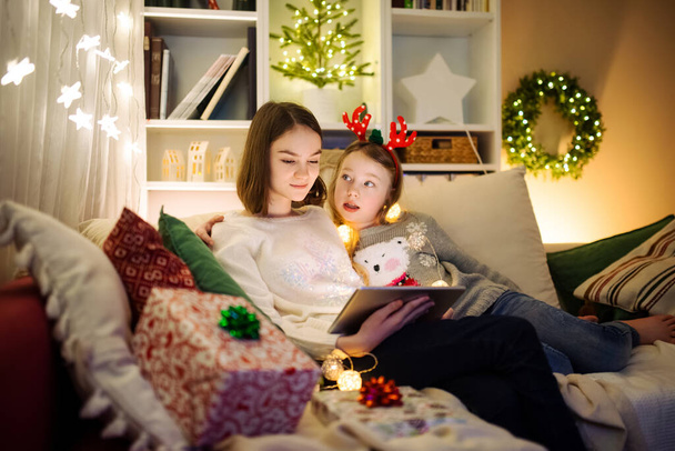 Two cute young sisters using a tablet pc at home in warm and cozy living room at Christmas. Family having online video call on Xmas eve. Winter evening at home with family and kids. - 写真・画像