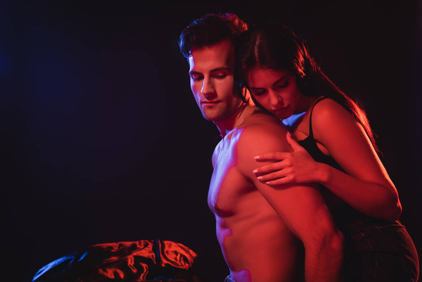 sexy brunette woman hugging shirtless muscular man in red lighting isolated on black - Foto, immagini