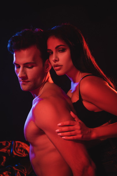 passionate brunette woman looking at camera while embracing shirtless man in red lighting isolated on black - Photo, Image