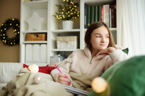 Beautiful teenage girl studying or sketching snuggled up on the sofa in a cozy living room at Christmas. Cute child spending time at home during winter break. - Foto, afbeelding