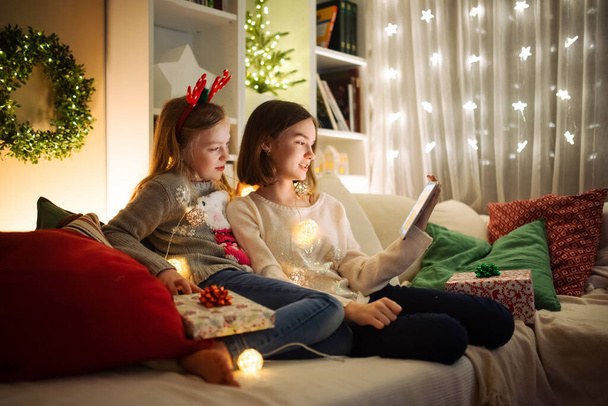 Two cute young sisters using a tablet pc at home in warm and cozy living room at Christmas. Family having online video call on Xmas eve. Winter evening at home with family and kids. - Foto, Imagem