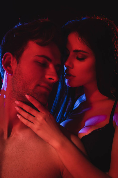 passionate woman in bra touching face of sexy man in red lighting isolated on black - 写真・画像