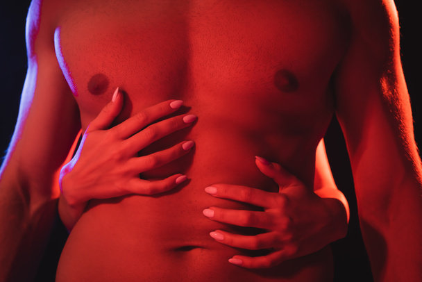 bijgesneden beeld van vrouw knuffelen gespierde romp van jonge man in rood licht geïsoleerd op zwart - Foto, afbeelding