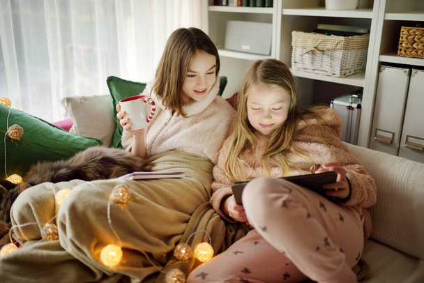 Two young sisters snuggling up on the sofa in a cozy living room at Christmas. Cute children using a tablet at home during winter break. Kids reading a book in comfy blanket. Celebrating Xmas at home - Fotó, kép
