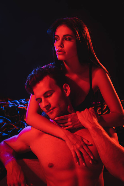 young brunette woman hugging shirtless muscular man in red lighting isolated on black - Valokuva, kuva