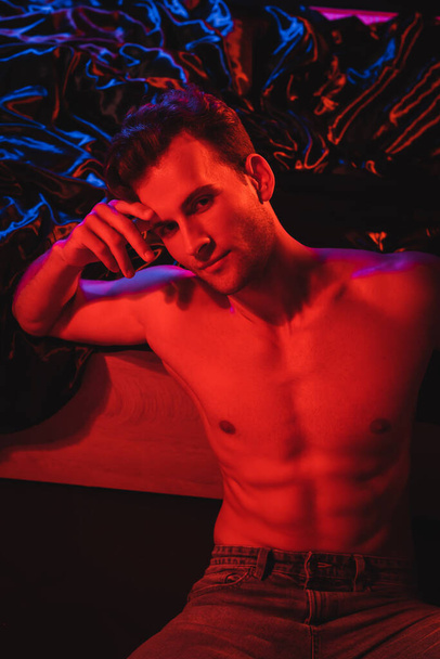 sexy shirtless man looking at camera near black bedding in red lighting - Photo, Image