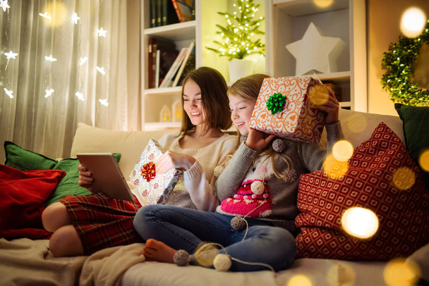 Two cute young sisters using a tablet pc at home in warm and cozy living room at Christmas. Family having online video call on Xmas eve. Winter evening at home with family and kids. - Valokuva, kuva