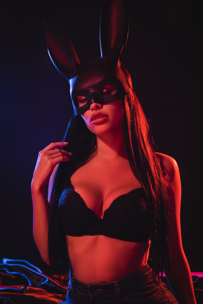 mujer sensual en sujetador y máscara de conejito mirando a la cámara sobre fondo negro con iluminación - Foto, Imagen