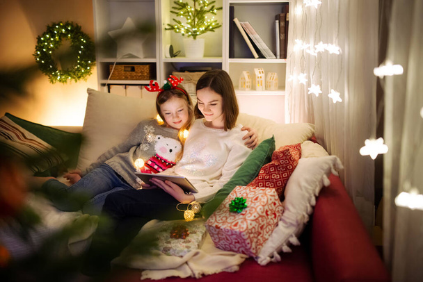 Two cute young sisters using a tablet pc at home in warm and cozy living room at Christmas. Family having online video call on Xmas eve. Winter evening at home with family and kids. - Zdjęcie, obraz