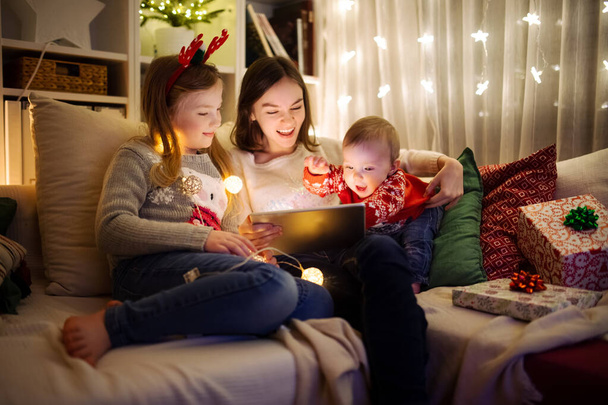 Two girls and a baby boy using a tablet pc at home in warm and cozy living room at Christmas. Family having online video call on Xmas eve. Winter evening at home with family and kids. - Fotografie, Obrázek