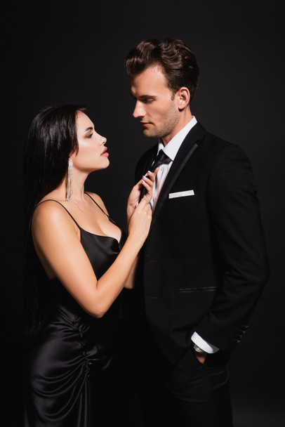 brunette woman seducing man in elegant suit and touching his tie isolated on black - Foto, imagen