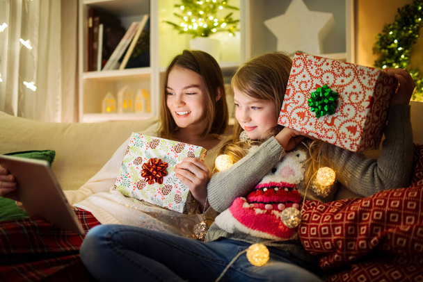 Two cute young sisters using a tablet pc at home in warm and cozy living room at Christmas. Family having online video call on Xmas eve. Winter evening at home with family and kids. - Foto, imagen