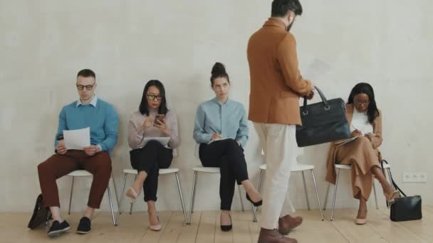 Foto media de grupo de personas sentadas en silla en fila y esperando una entrevista de trabajo. Están leyendo formularios de solicitud y utilizando el teléfono móvil - Metraje, vídeo