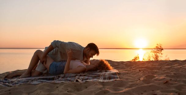 Passionate young couple kissing while lying together on the beach at sunset - Фото, изображение