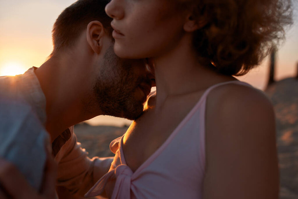 Genç bir adamın köprücük kemiğinden bir kadını öperken görüntüsü. Çiftler gün batımında sahilde eğleniyorlar. - Fotoğraf, Görsel