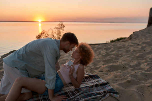 Beautiful young couple lying on the beach at sunset, ready for making love. Woman is holding a condom - Foto, afbeelding