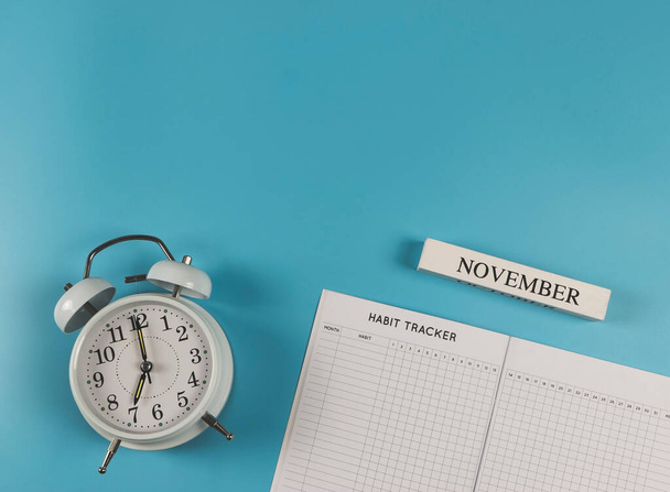 Bovenaanzicht of platte lay-of gewoonte tracker boek, witte vintage wekker en houten kalender november op blauwe achtergrond. - Foto, afbeelding