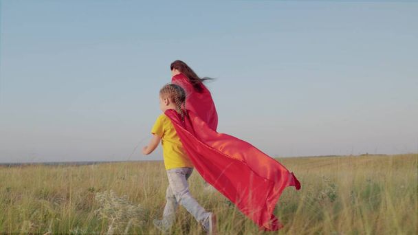 Mother and child run together across green field in red raincoats. Kid dreams of becoming superhero. Happy family mom and daughter play together in super heroes. Childrens games dreams. Slow motion. - Foto, Imagen