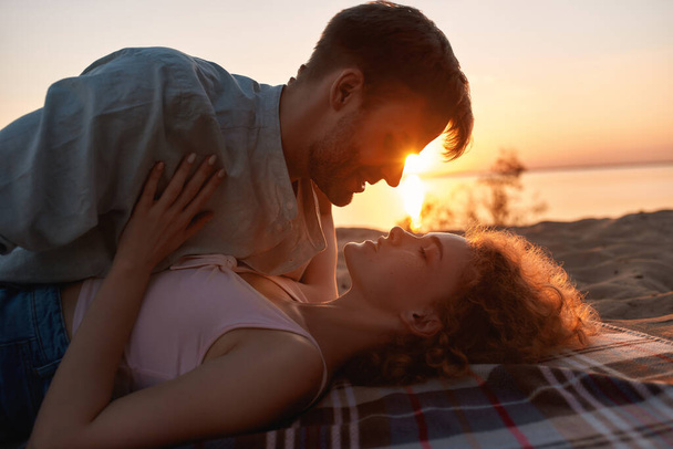 Passionate young couple kissing on the shore with the sun behind them, lying together on the beach at sunset - Foto, imagen