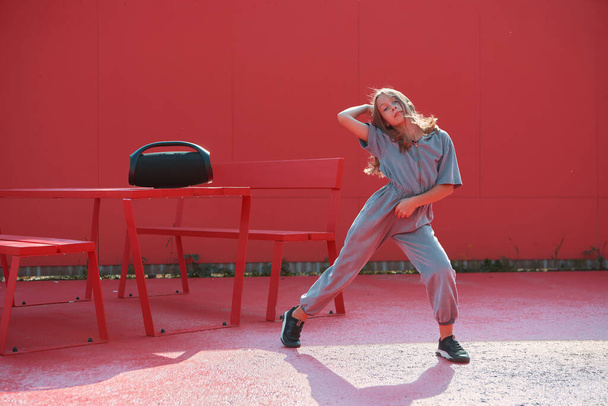 Active teen girl dancing hip hop with wireless outdoor speaker outside on red background. Dance school advertisement - Foto, imagen