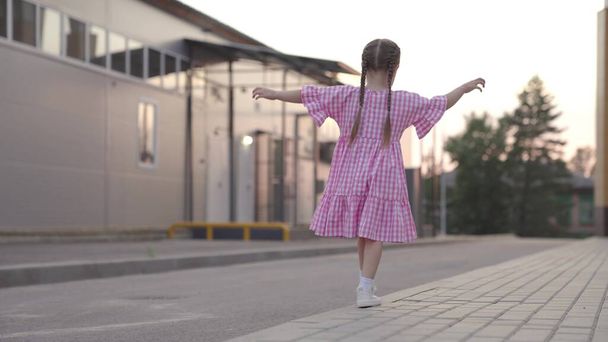 маленькая девочка подняла руки и держит равновесие, детские игры на улице, ребенок веселится, ребенок гуляет по бардюру в городе, активные детские развлечения во дворе - Фото, изображение