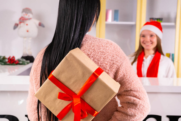 Back view photo of a young brunette woman holding the present box with the red ribbon behind the back to give it to her colleague in a Santa hat and red scarf.  - Foto, Bild