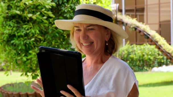 帽子の熱心なシニア女性ブロンドの実業家は、ビジネスの仕事のためのタブレットPC 、デジタルタブレットを使用するか、彼女自身の緑の庭で勉強します。50歳から55歳の女性。彼ら自身のビジネスとトレーニングの概念 - 映像、動画