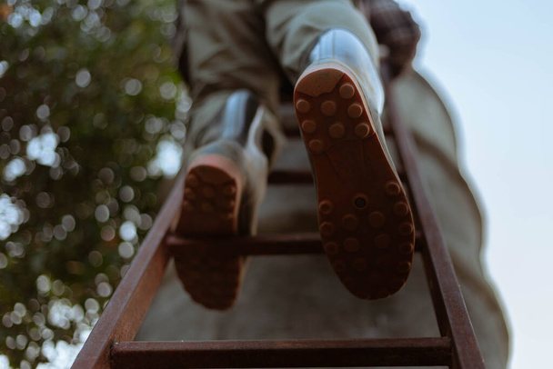 Concepto de jardinería un agricultor masculino que usa botas grandes que suben en una escalera de madera a un tanque de agua de cemento. - Foto, Imagen