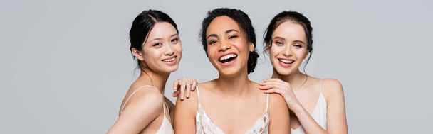 Cheerful multicultural women looking at camera isolated on grey, banner  - Photo, Image