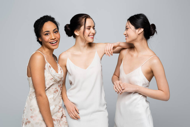 Positive multiethnic women in satin dresses standing isolated on grey  - Foto, immagini