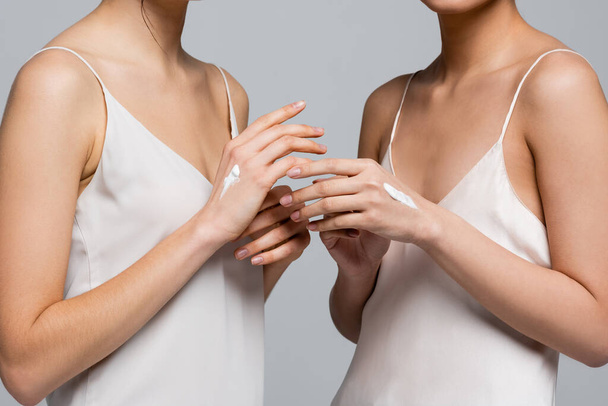 Обрезанный вид молодых женщин в атласных платьях и кремах на руках, изолированных на сером  - Фото, изображение
