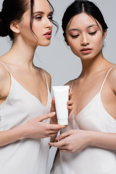 Pretty multiethnic women in silk dresses holding cream in tube isolated on grey  - Foto, afbeelding