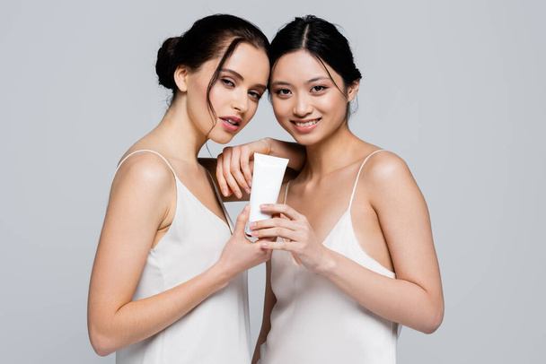 Smiling asian woman in satin dress holding cream near friend isolated on grey  - Foto, Bild