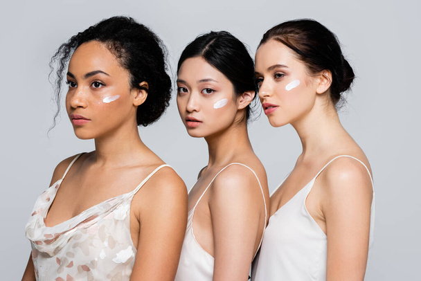 Mujeres jóvenes interraciales con crema cosmética en las caras aisladas en gris  - Foto, imagen