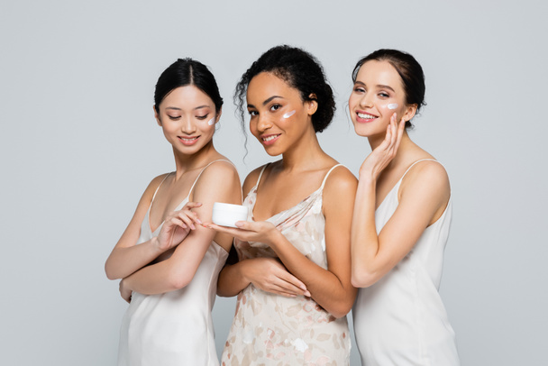 Positive african american woman holding container with cream near interracial friends isolated on grey  - 写真・画像