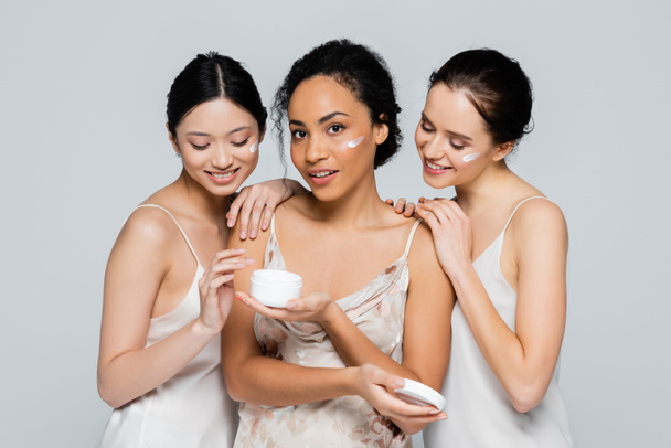Interracial women hugging african american friend with cosmetic cream isolated on grey  - Fotó, kép