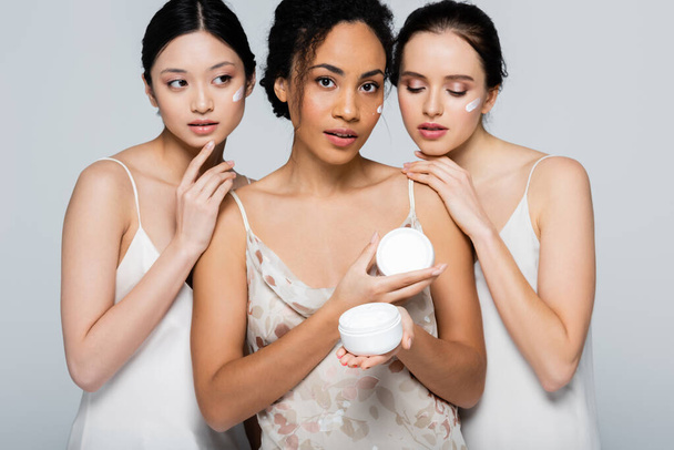 African american woman with cosmetic cream looking at camera near multicultural friends isolated on grey  - Photo, Image