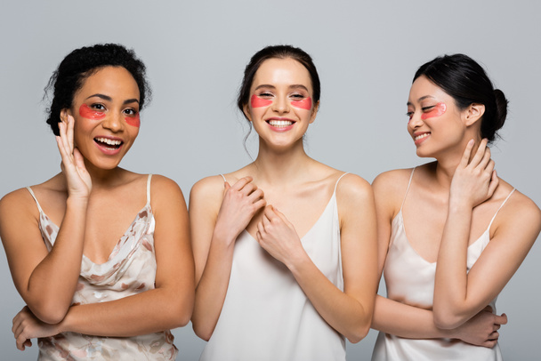 gelukkig interraciale vrouwen met ogen patches staan geïsoleerd op grijs  - Foto, afbeelding