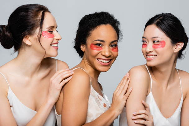 Glückliche interrassische Frauen mit Augenklappen, die einander berühren, isoliert auf grau  - Foto, Bild