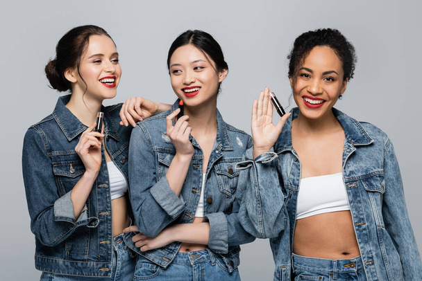 Smiling multiethnic women in denim jackets holding lipsticks isolated on grey  - 写真・画像