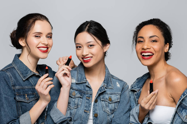 Smiling interracial friends holding red lipsticks isolated on grey  - Photo, Image